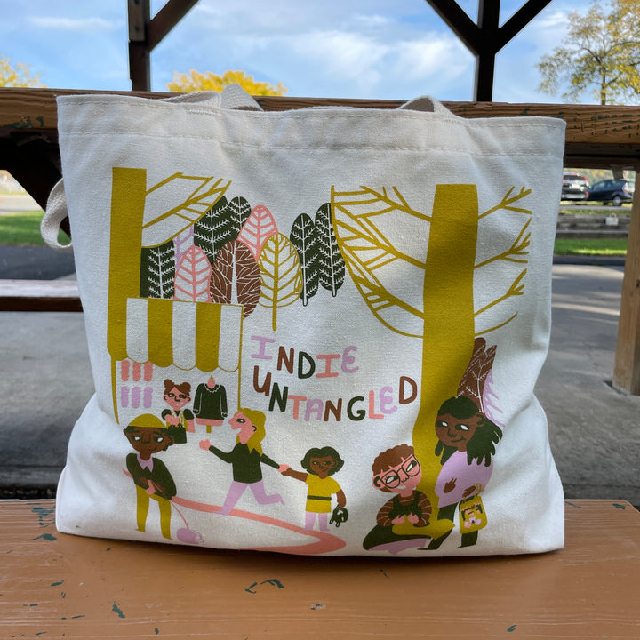 A cream tote bag with a gold, brown, pink, purple and gray illustration.