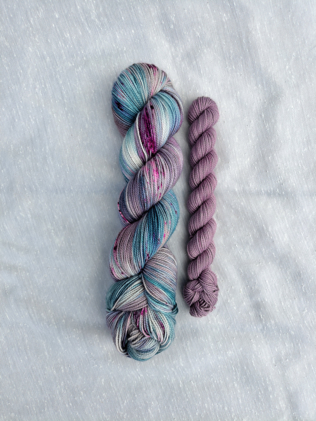 A full skein of blue, purple and green speckled yarn and a mini skein of pale purple semisolid yarn.