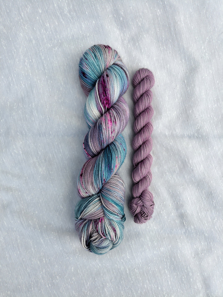 A full skein of blue, purple and green speckled yarn and a mini skein of pale purple semisolid yarn.