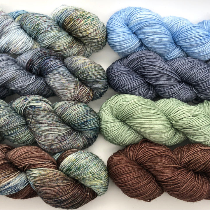 Eight skeins of yarn displayed in two neat rows. The colors vary and include soft shades of green, blue, gray, brown, and multi-colored speckled blends.