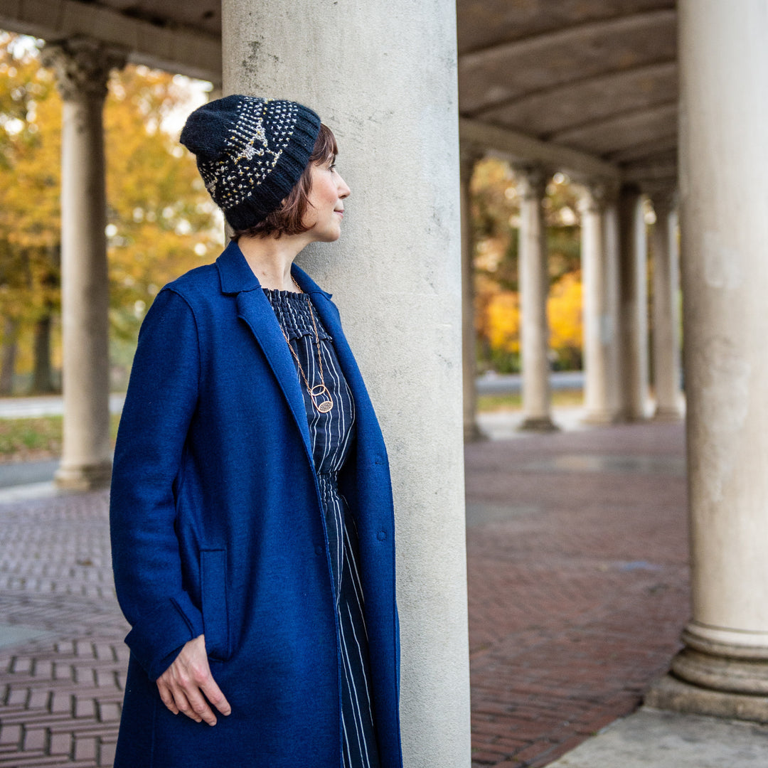 A white woman with short brown hair in a royal blue coat wears a navy beanie with gray in the pattern of a menorah.