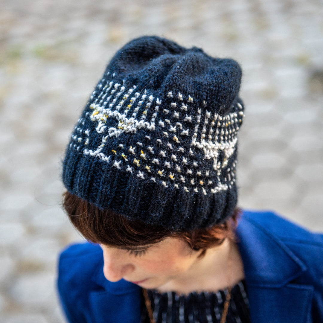 A white woman with short brown hair in a royal blue coat wears a navy beanie with gray in the pattern of a menorah.