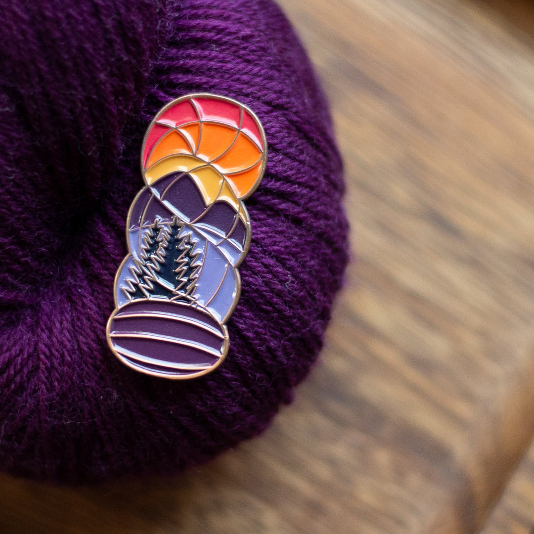 A pin in the shape of a hank of yarn with the image of an orange sunset over purple mountains.