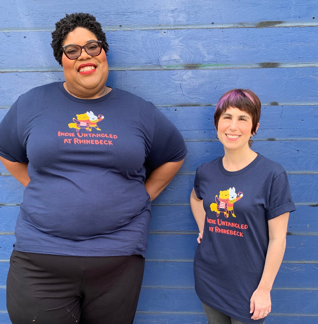 An African American, plus-sized woman with curly hair stands next to a petite Caucasian woman with a pixie cut. They’re wearing matching navy T-shirts.