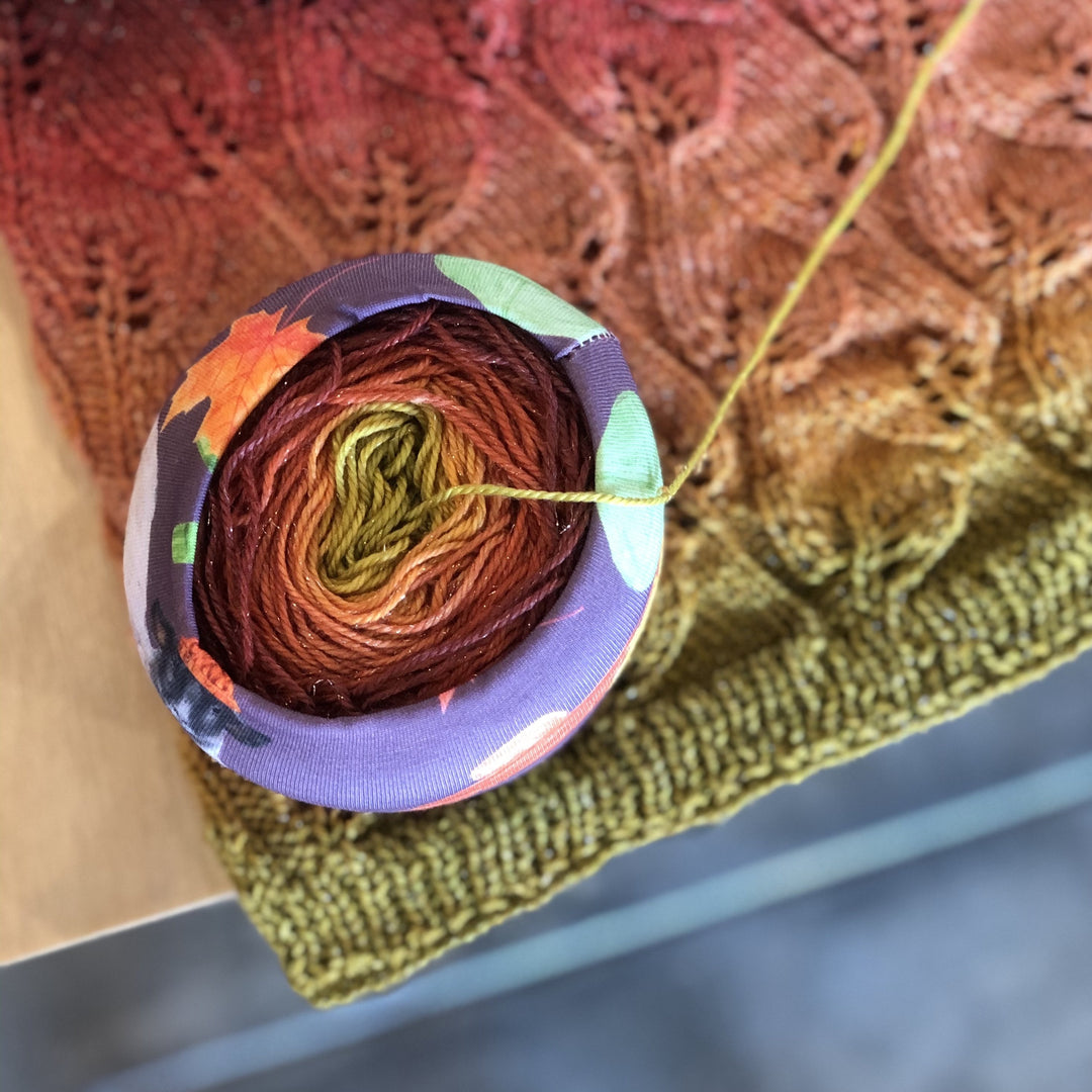 A purple, orange and green sleeve holding a cake of red to green ombre yarn.