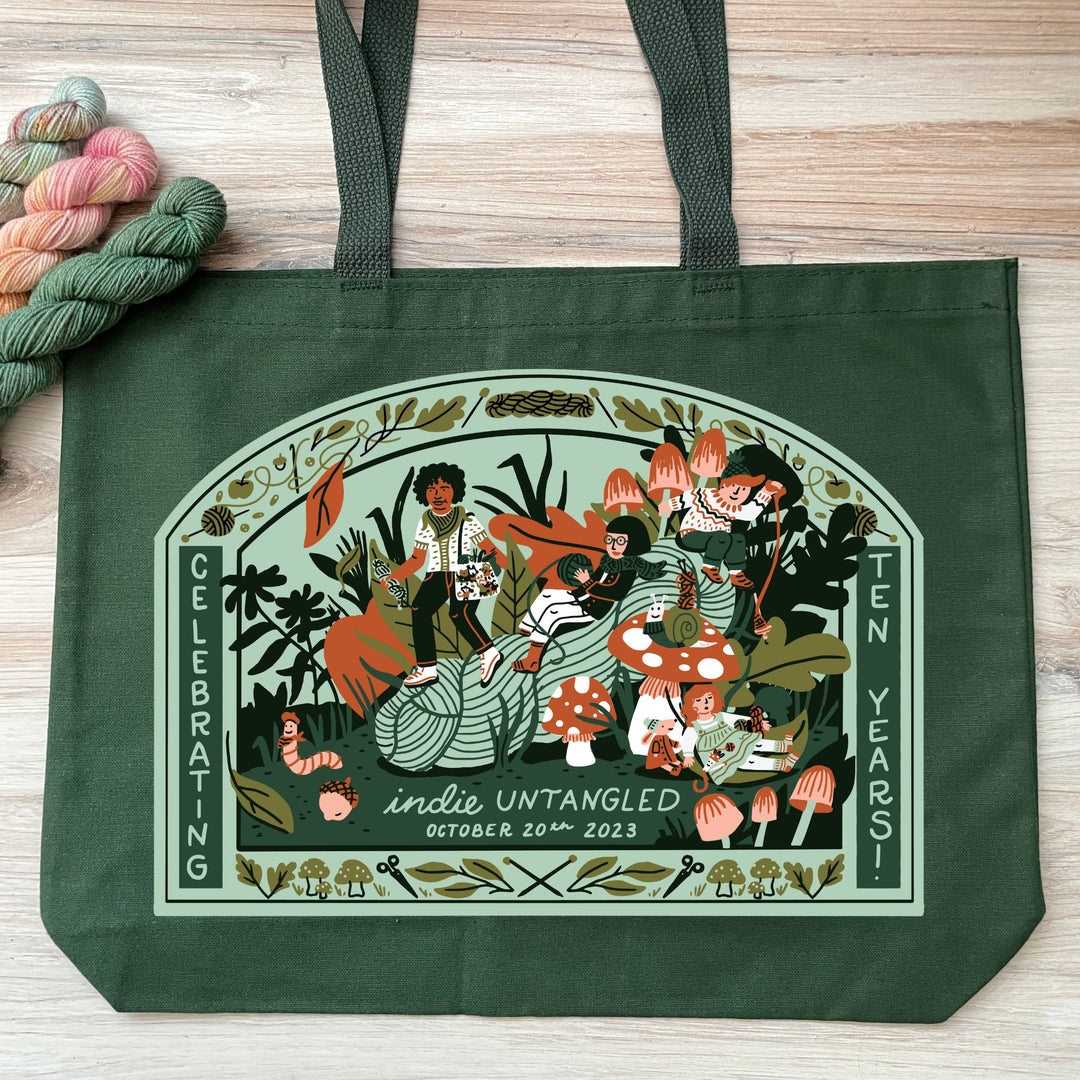 A green bag with an illustration in greens and oranges of a diverse group of people lounging and frolicking around a giant skein of yarn. The text reads Indie Untangled, October 20, 2023, Celebrating ten years!
