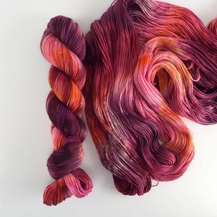 A single skein of yarn next to its unraveled strands. The yarn is hand-dyed with vibrant shades of magenta, plum, orange and pink, with subtle tonal variations. 