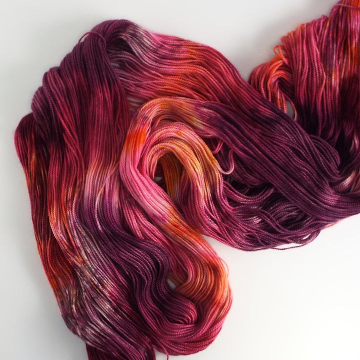 An unraveled skein of yarn hand-dyed with vibrant shades of magenta, plum, orange and pink, with subtle tonal variations. 