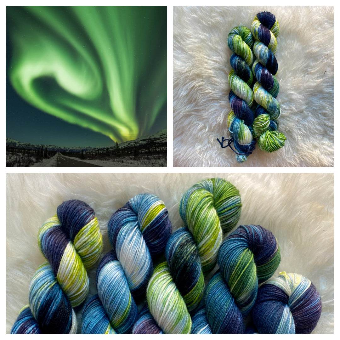 A wave of electric green light swirls in a blue sky above snow-covered mountains and spruce trees. Hand-dyed yarn in deep indigo, vibrant green and bright blue hues.