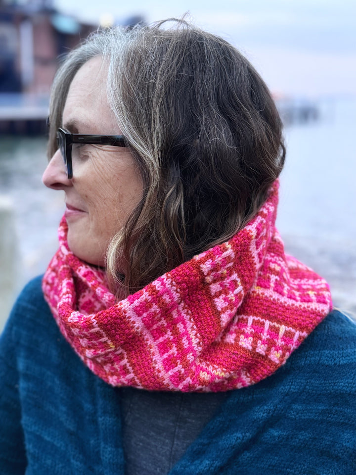 Side view of a light-skinned woman with glasses wearing a pink, fuchsia and orange colorwork cowl.