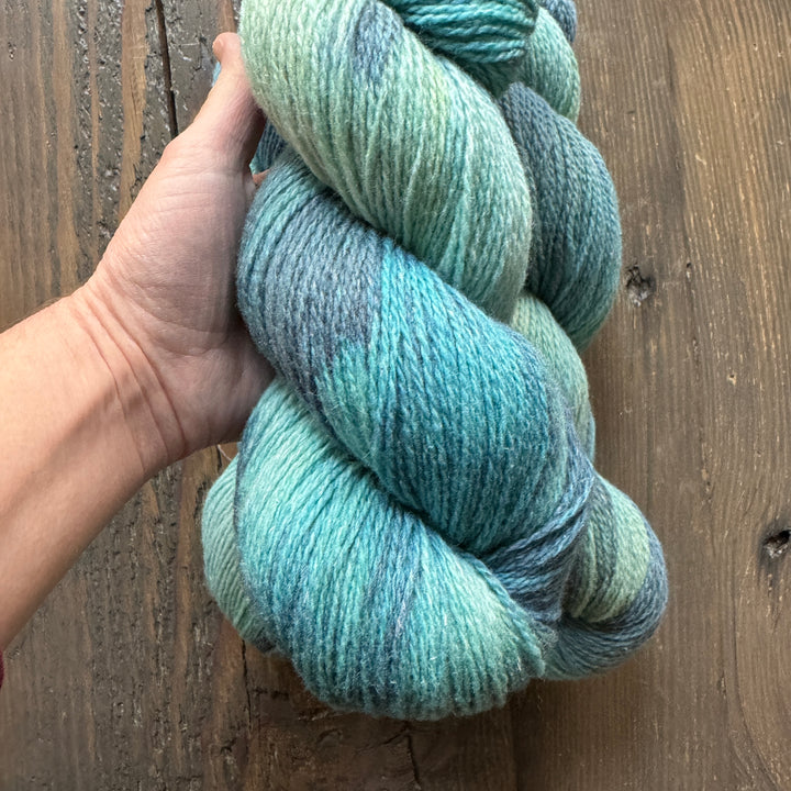 A light-skinned hand holds a skein of Skeins of light blue/green variegated yarn.