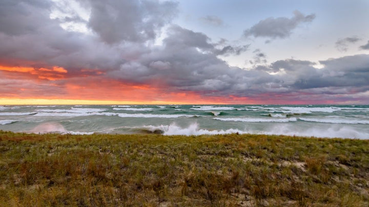 A lakeshore at sunset. The sky is filled with dramatic clouds, and the setting sun casts vibrant orange and red hues across the horizon. 
