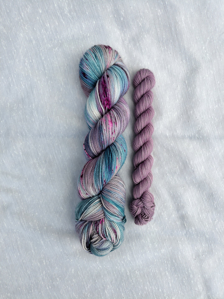  A full skein of blue, purple and green speckled yarn and a mini skein of pale purple semisolid yarn.