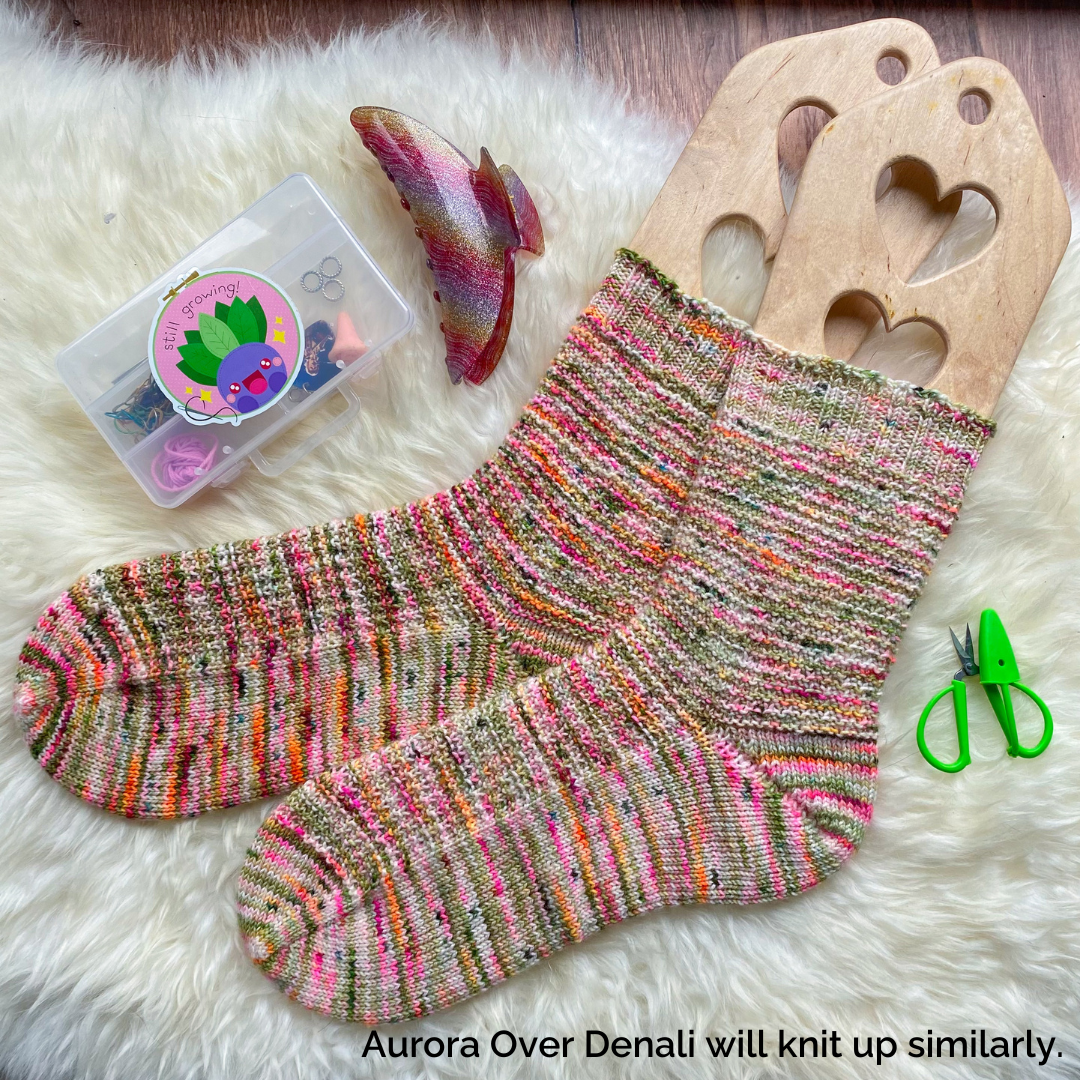 Hand knit socks in speckles of pink, orange and green on wooden blockers. The text Aurora Over Denali will knit up similarly.