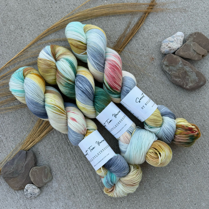 Skeins of hand-dyed yarn in a soft, variegated color palette with shades of cream, teal, mustard, and specks of pink and gray. 