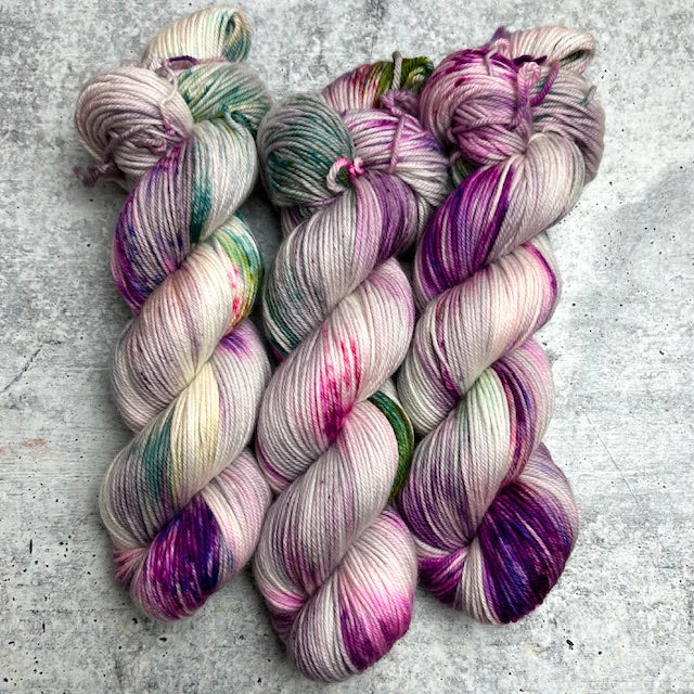 Three skeins of hand-dyed yarn laid side by side. The yarn features a blend of light gray, white, purple, pink, and green colors.