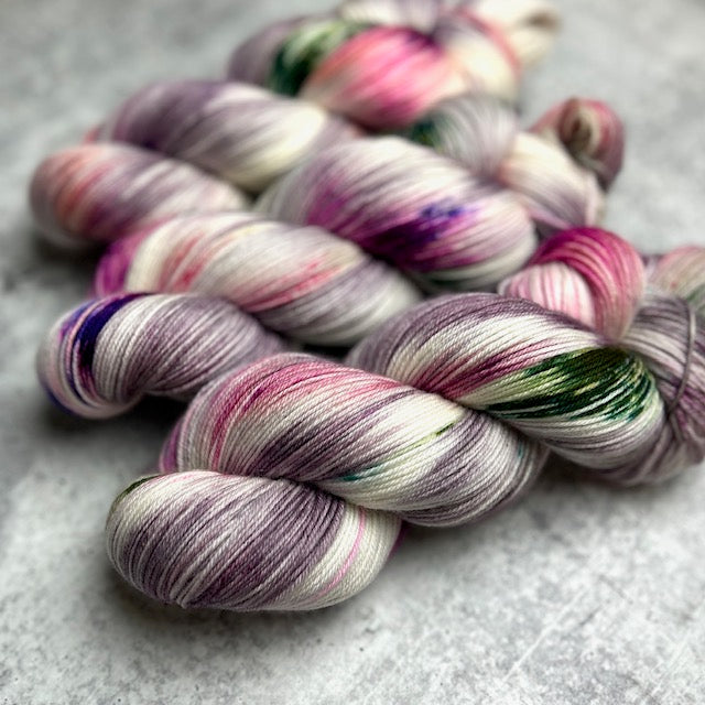 A close-up of skeins of hand-dyed yarn. The yarn is predominantly white and light gray with splashes of vibrant colors like purple, pink, and green.