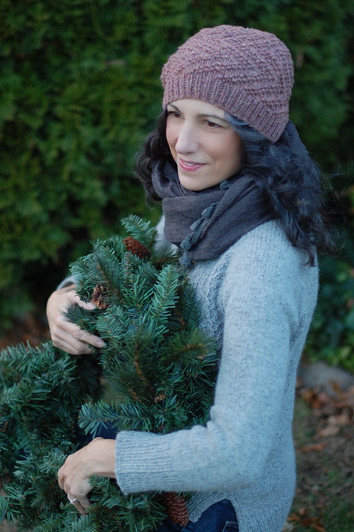 A white woman wears a pink beanie with textured arrows and carries a green wreath.