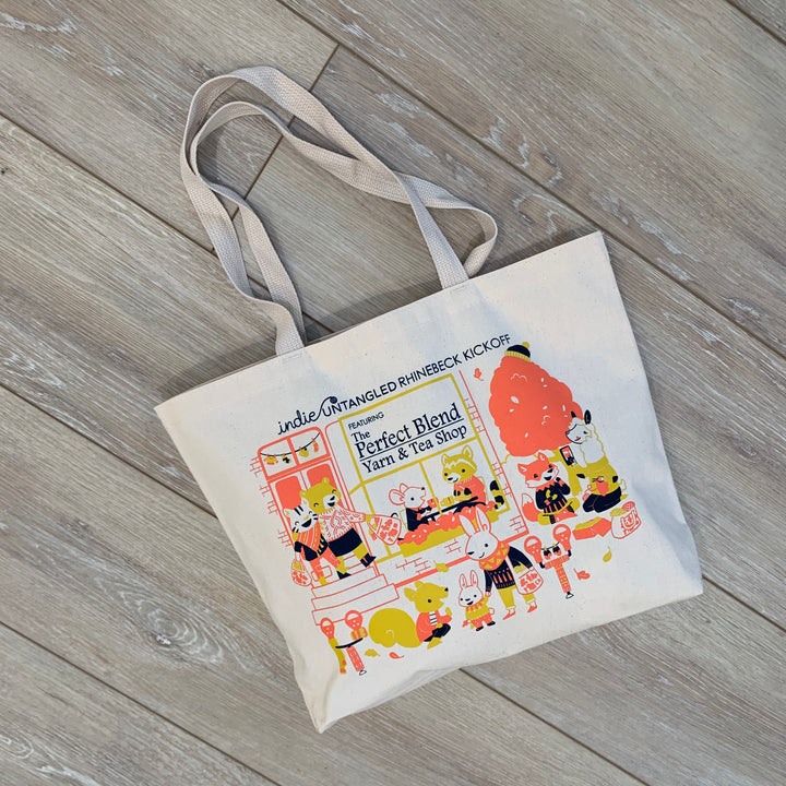 A white cotton tote bag with a coral, yellow and blue illustration of animals outside of a yarn shop. 
