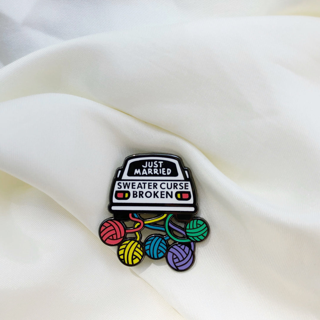 An enamel pin of white car with rainbow yarn balls streaming behind it and the words JUST MARRIED SWEATER CURSE BROKEN.