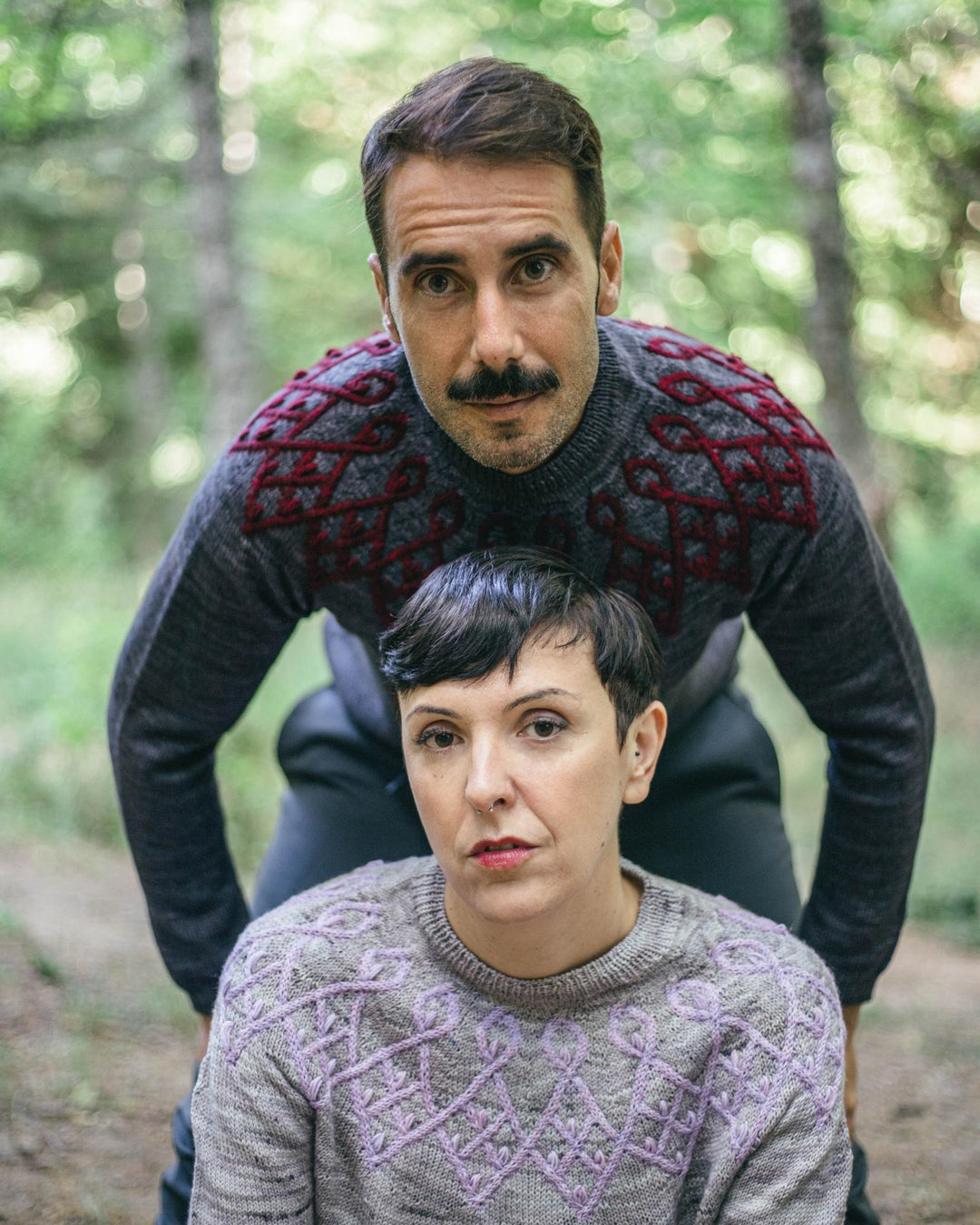 A man and a woman wearing colorwork sweaters. His is dark gray and red and hers is light gray and lilac.