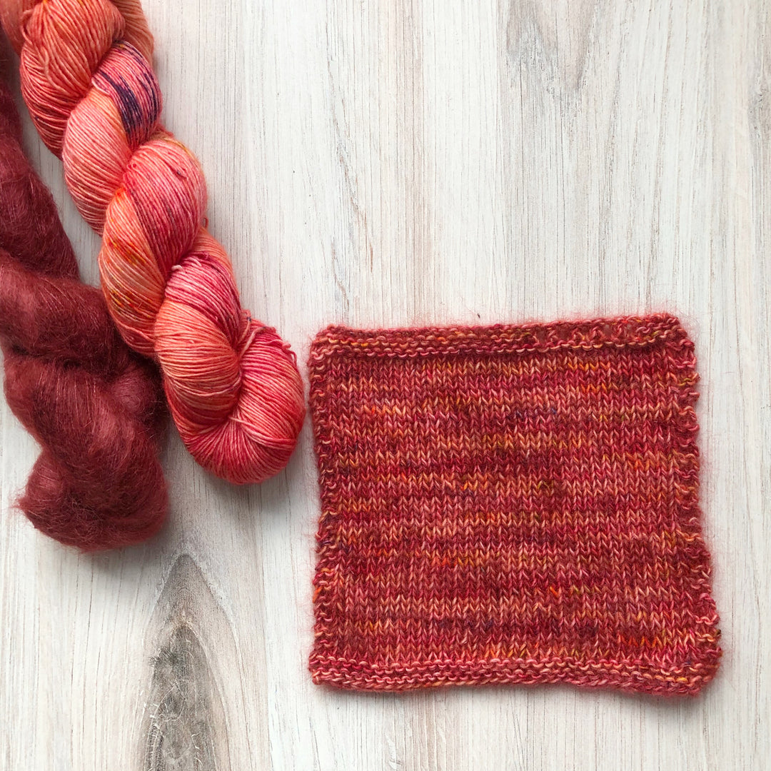 A swatch of knit fabric in red and orange next to skeins of purple mohair and red-orange speckled yarn.