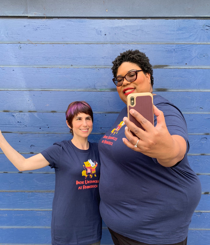 An African American, plus-sized woman with curly hair stands next to a petite Caucasian woman with a pixie cut. They’re wearing matching navy T-shirts.