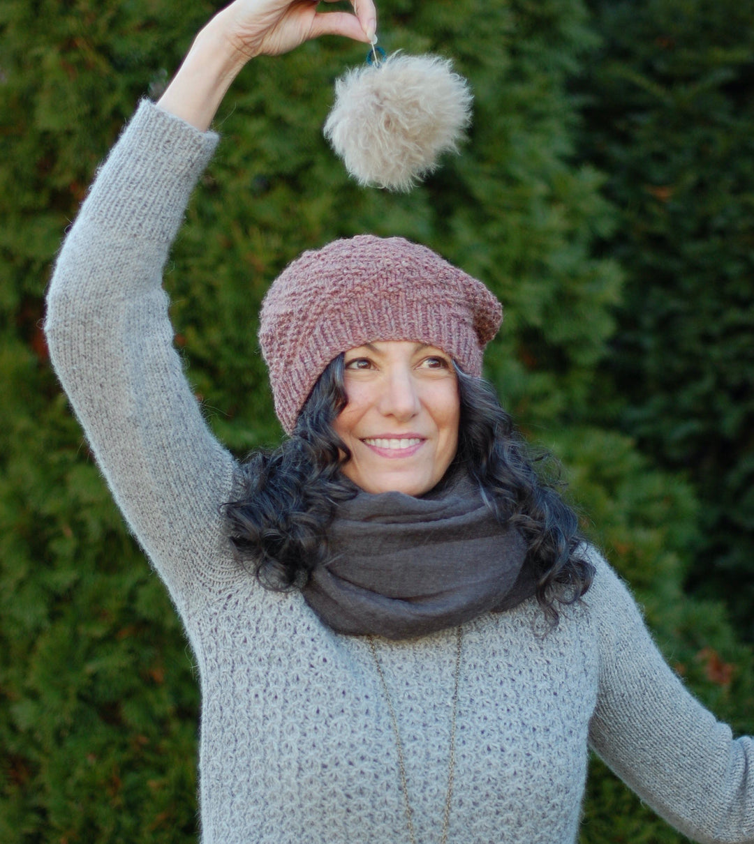 A white woman wears a pink textured beanie and holds a fluffy pompom over her head.