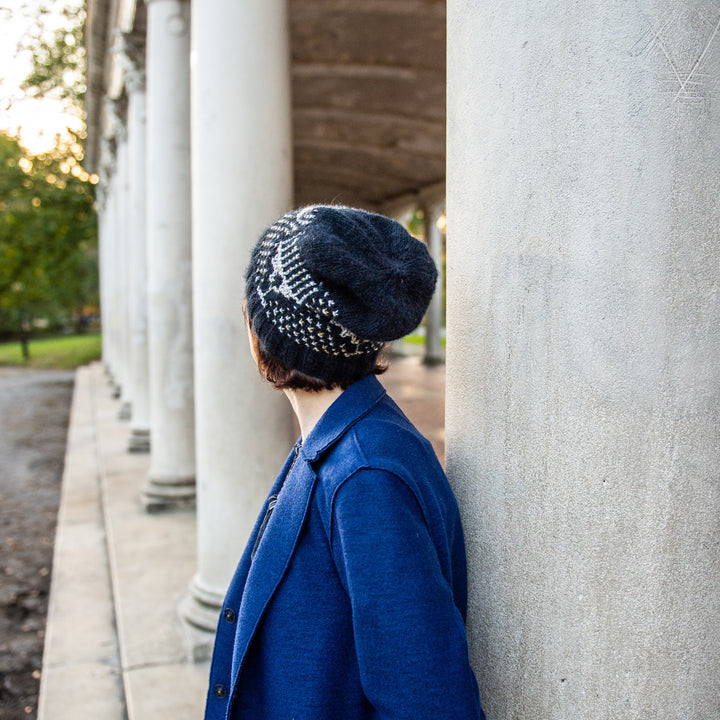 A white woman with short brown hair in a royal blue coat wears a navy beanie with gray in the pattern of a menorah. 