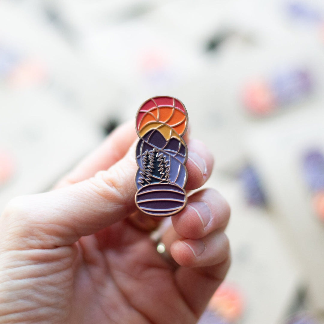 A pin in the shape of a hank of yarn with the image of an orange sunset over purple mountains. 