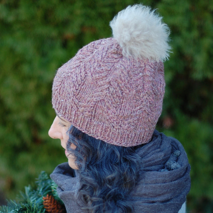 A white woman wears a pink cabled beanie with a fluffy white pompom. 