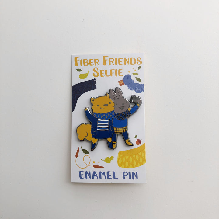 A blue enamel pin of a squirrel and bunny taking a selfie with a phone. 