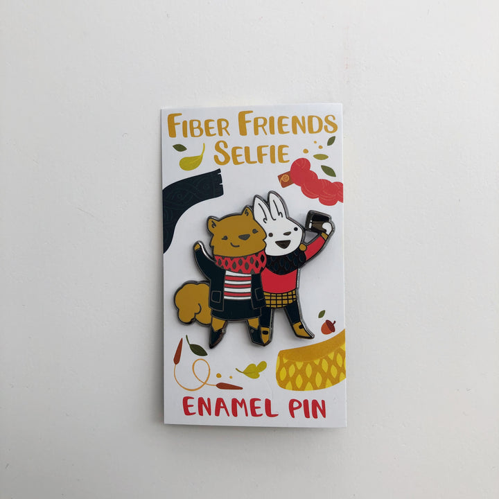 A coral enamel pin of a squirrel and bunny taking a selfie with a phone. 