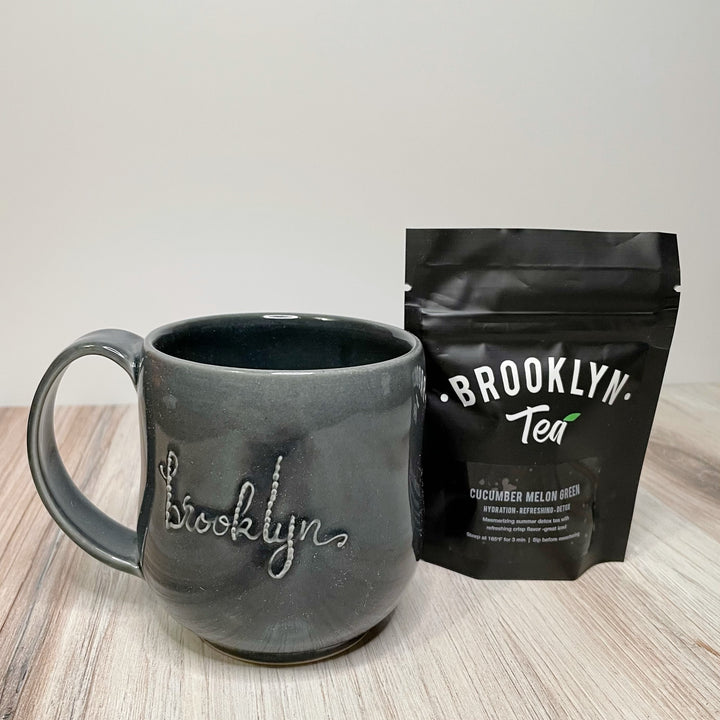 A gray curved mug with the word Brooklyn in script and a black pouch that reads Brooklyn Tea, Cucumber Melon Green.