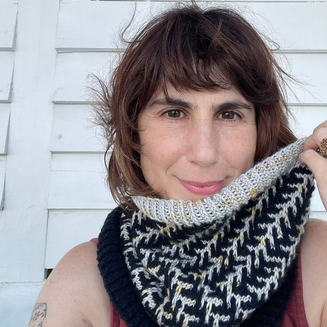 A white woman with wavy brown hair wears a cowl in dark navy with pine boughs in silver yarn with gold speckles.