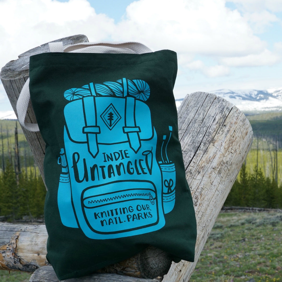 A green tote bag with the illustration of a backpack and the words Indie Untangled Knitting Our Natl Parks.