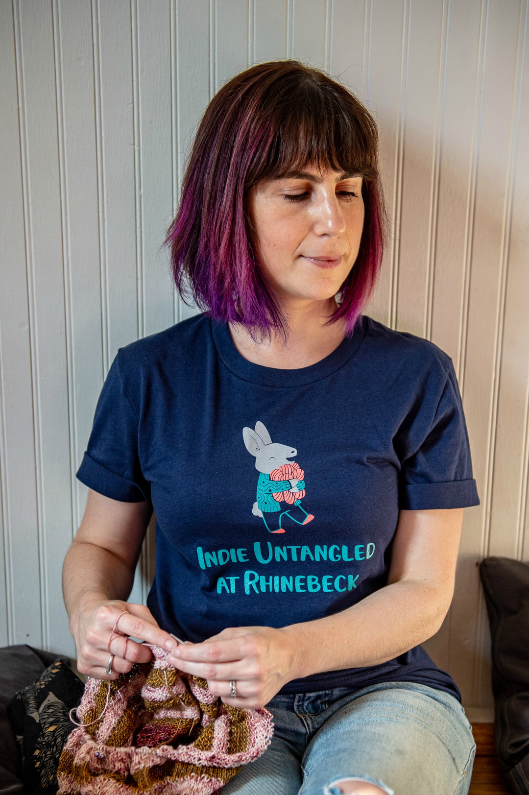 A white woman wears a navy t-shirt with an illustration of a bunny clutching coral yarn and the words Indie Untangled At Rhinebeck.