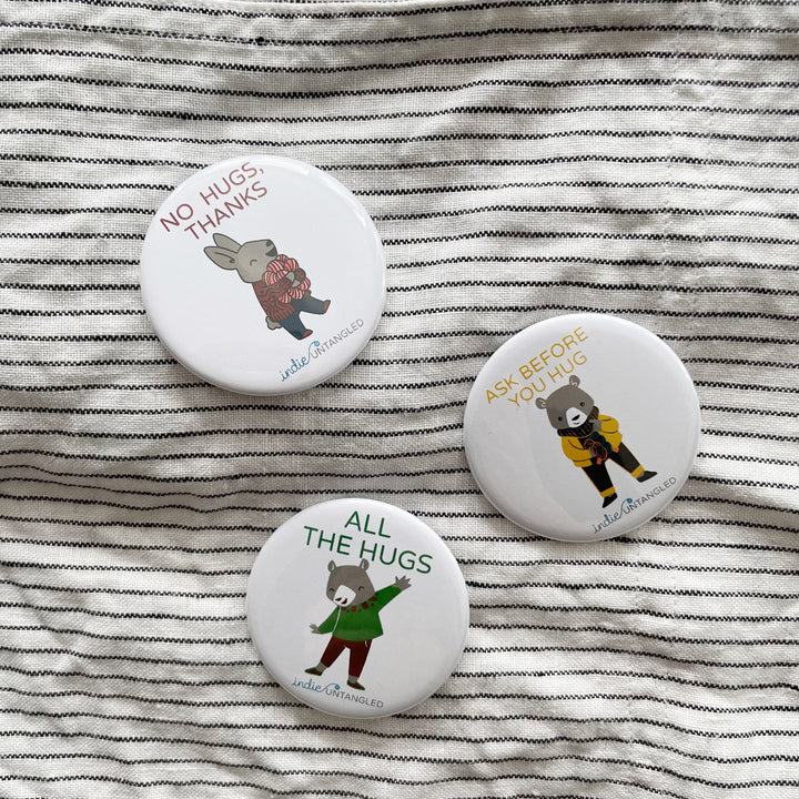 Round buttons with a rabbit wearing a red sweater and the words No hugs, thanks, a bear wearing a green sweater and the words All the hugs and a bear wearing a yellow sweater and the words Ask before you hug.