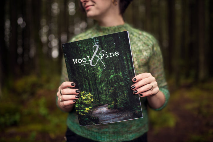 A white woman holds a book with a photo of a forest that reads Wool & Pine.