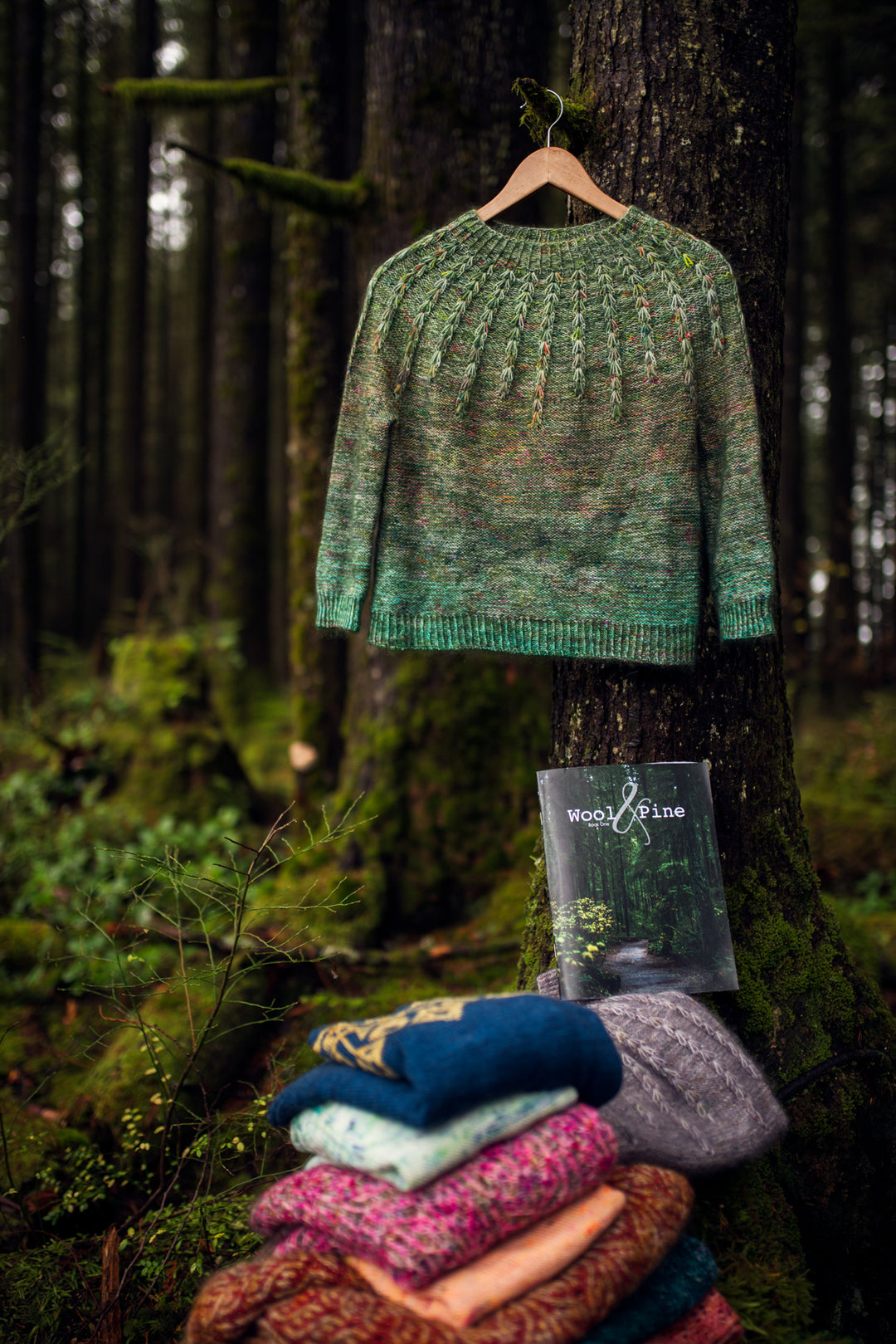 A green ombre sweater hangs from a tree branch over a pile of colorful knitwear.