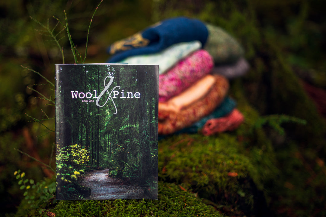 A book with a photo of a forest that reads Wool & Pine in front of an out-of-focus pile of colorful knitwear.