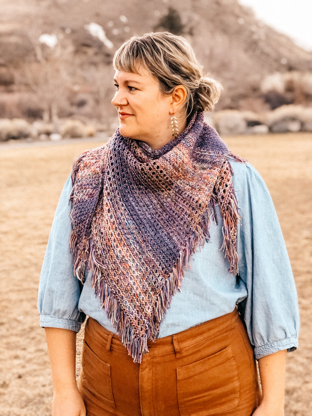 A textured and eyelet shawl with fringe in orange, purple and blue faded yarn, worn by a light-skinned woman standing in front of a mountain.