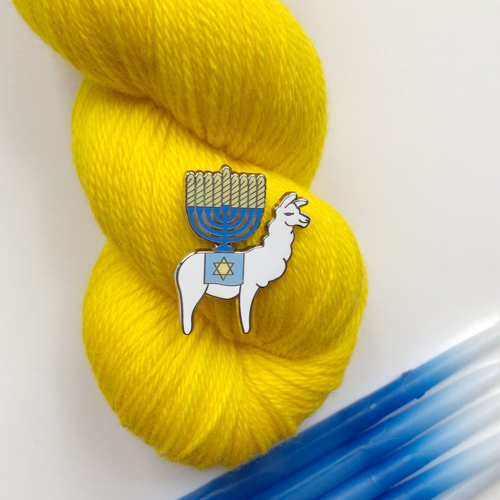 An enamel pin of a white llama with a blue and yellow menorah on its back on a skein of yellow yarn