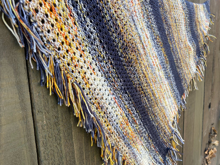 A textured and eyelet shawl with fringe in gray, orange, brown, yellow and white yarn faded together. 