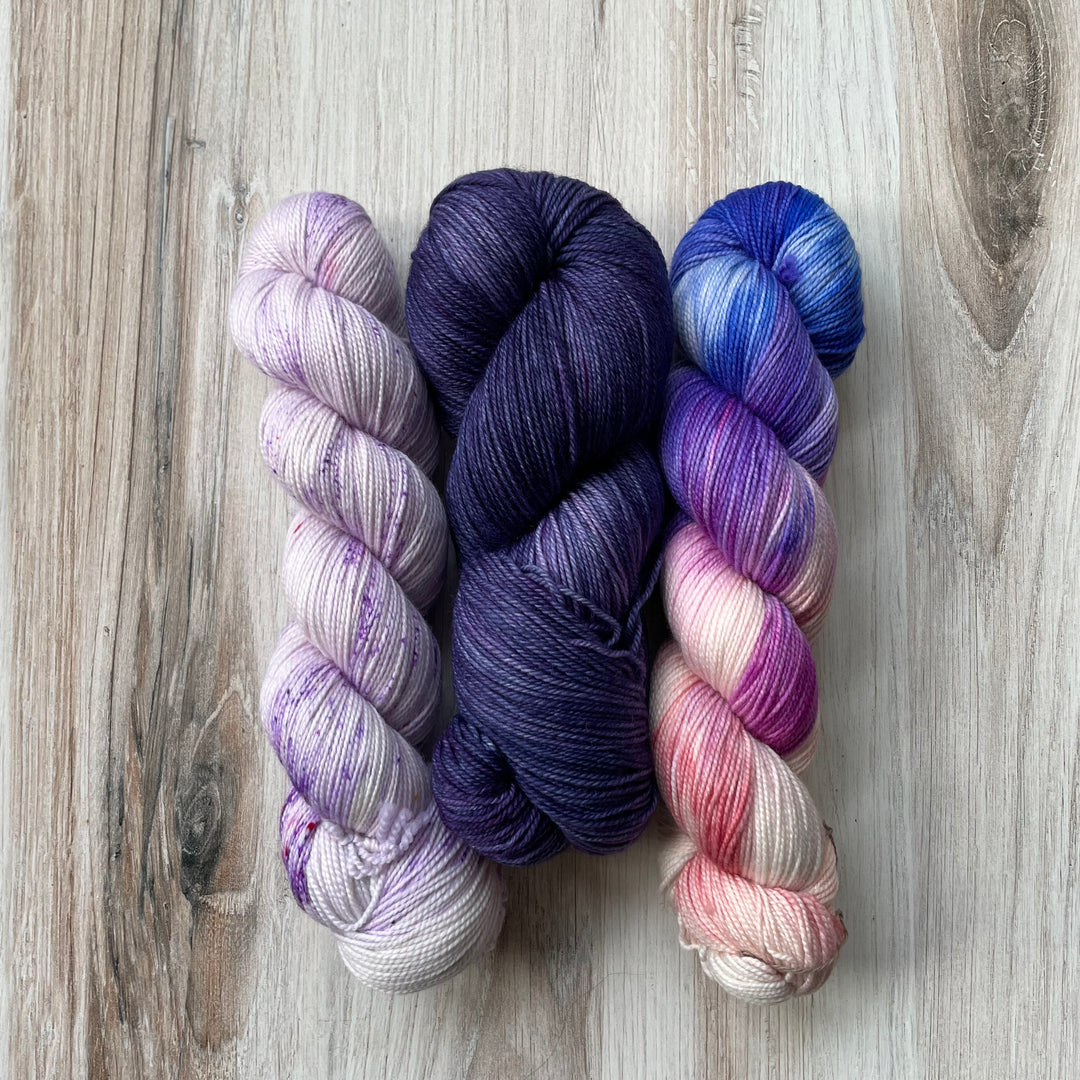 Three skeins of yarn in purple speckle, solid purple and variegated purple, blue and pink. 