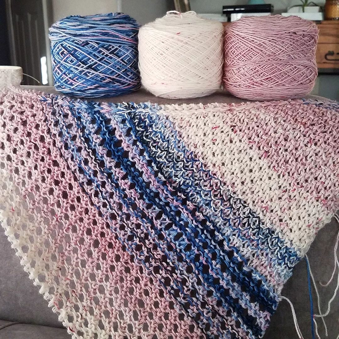 A textured and eyelet shawl on the needles in shades of pale pink with faint pink speckles, white speckled with pink and blue and pink variegated yarn, below yarn cakes in the same colors. 