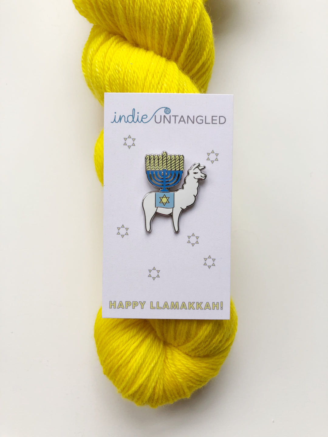 An enamel pin of a white llama with a blue and yellow menorah on its back on a skein of yellow yarn.  