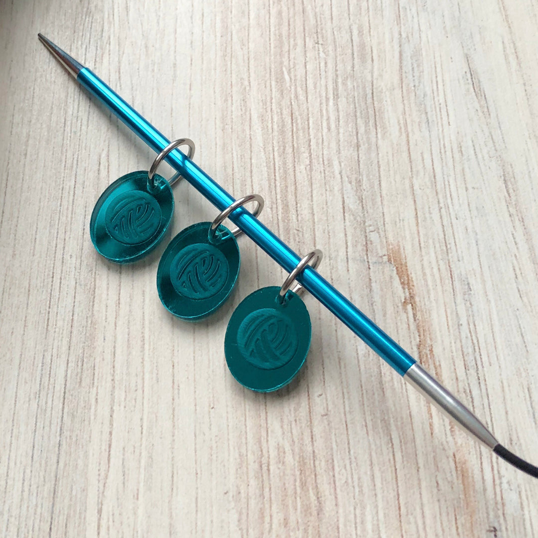 Three teal acrylic stitch markers with a yarn ball image on a blue knitting needle.