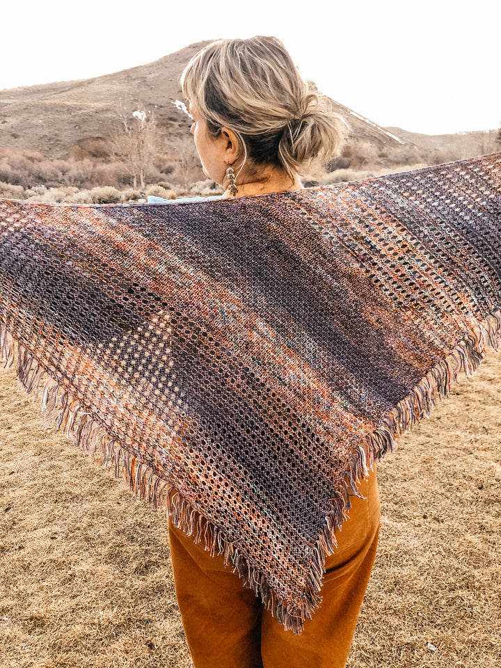 A textured and eyelet shawl with fringe in orange, purple and blue faded yarn, worn by a light-skinned woman standing in front of a mountain.