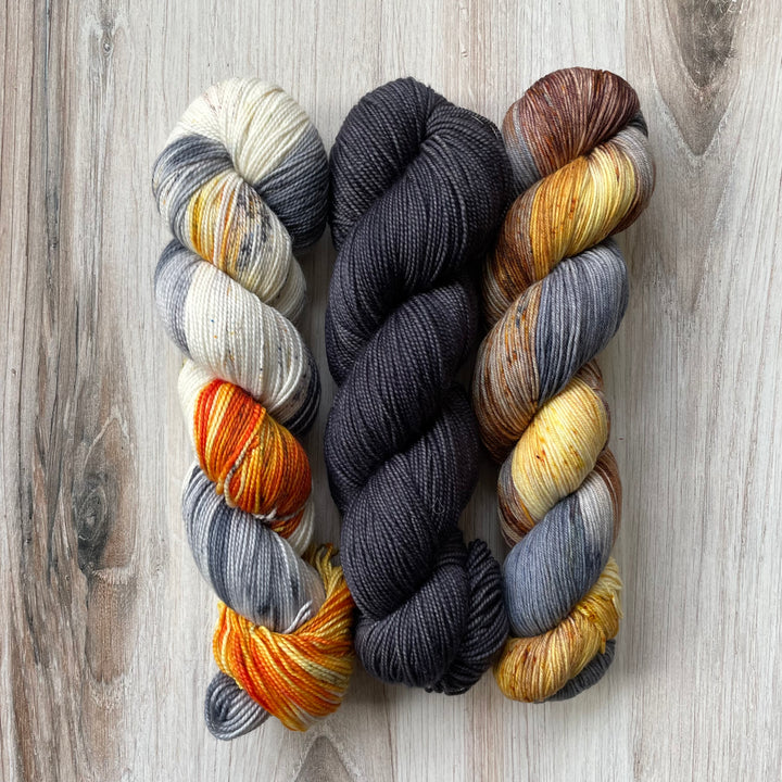 Three skeins of yarn in orange and gray speckle, solid gray and variegated gray, brown and yellow. 